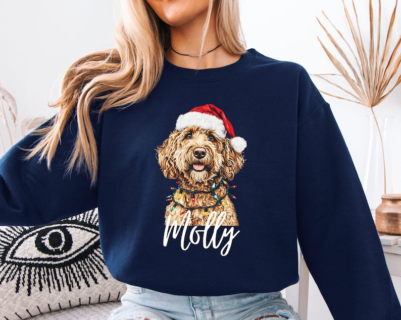 Pu&ograve; includere: Felpa blu navy con un cane golden doodle con cappello da Babbo Natale e luci. Il nome del cane, "Molly", &egrave; scritto in bianco. La felpa &egrave; a girocollo.