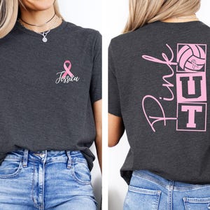 May include: Dark gray t-shirt with a pink ribbon and the name "Jessica" printed on the front. The back of the shirt features the word "Pink" in cursive, along with a volleyball and the letters "UT" in pink.