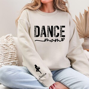 May include: A cream-colored sweatshirt with the text "Dance Mom" in black with a leopard print pattern. The sweatshirt has a black silhouette of a dancer on the left sleeve with the name "Jessica" underneath.