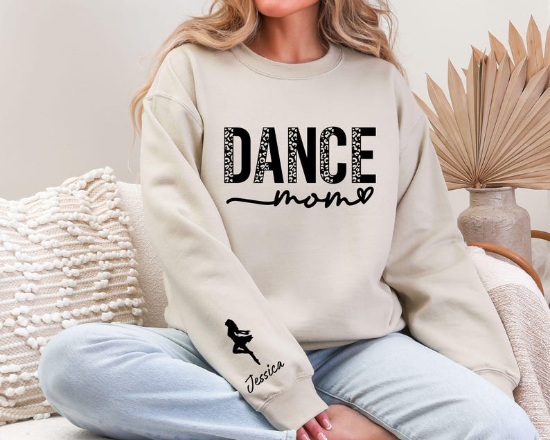 May include: A cream-colored sweatshirt with the text "Dance Mom" in black with a leopard print pattern. The sweatshirt has a black silhouette of a dancer on the left sleeve with the name "Jessica" underneath.