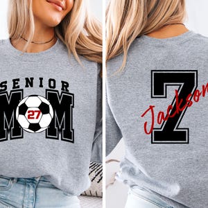 May include: A heather gray crewneck sweatshirt featuring a front graphic with the text SENIOR MOM and a soccer ball with the number 27. The back of the sweatshirt displays a large black number 7 with the name Jackson written in red script.