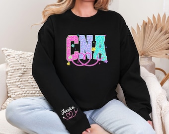 以下が含まれることがあります： ピンク、青、緑の「CNA」の文字がヒョウ柄でデザインされた黒いスウェットシャツ。 ピンクの聴診器のデザインと「Jessica」という名前が袖にあります。 クルーネックのスウェットシャツです。