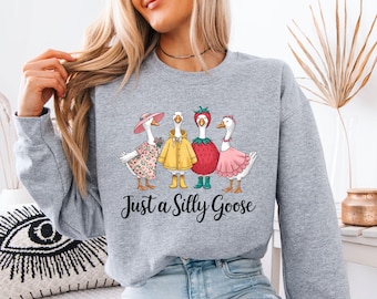 May include: A gray sweatshirt featuring a whimsical illustration of four geese dressed in various outfits. The geese are wearing a floral dress, a yellow raincoat, a strawberry costume, and a pink dress. The text "Just a Silly Goose" is below the illustration.