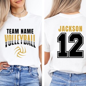 May include: White t-shirt with a volleyball design. The front features "TEAM NAME VOLLEYBALL" in black and gold lettering, with a volleyball graphic. The back displays "JACKSON 12" in gold and black.