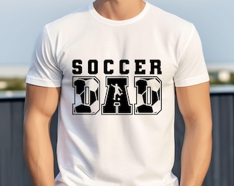 Può includere: T-shirt bianca con la scritta "SOCCER DAD" in grassetto nero. Le lettere "D" e "A" sono disegnate con un pallone da calcio e una silhouette di calciatore. Una maglietta casual per tutti i giorni.
