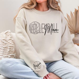 May include: Beige sweatshirt with the words "Volleyball Mom" in black script and a volleyball graphic. The sleeve features a volleyball and the name "Jessica". The sweatshirt is a crew neck style.