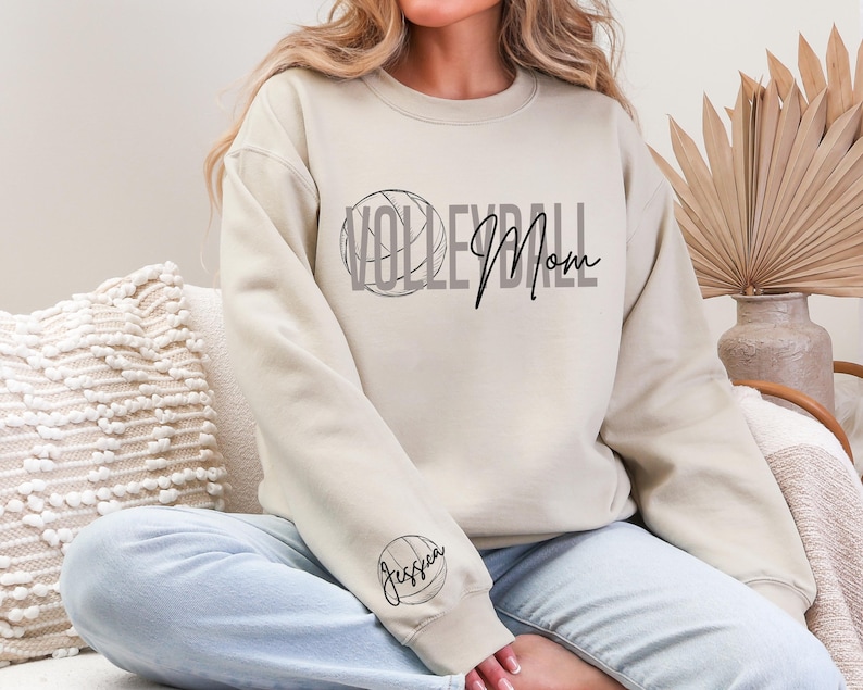 May include: Beige sweatshirt with the words "Volleyball Mom" in black script and a volleyball graphic. The sleeve features a volleyball and the name "Jessica". The sweatshirt is a crew neck style.