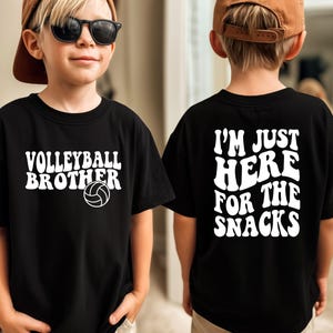 May include: Black t-shirt with white text that reads "Volleyball Brother" and a volleyball graphic on the front. The back of the shirt says "I'm Just Here For The Snacks" in white text. The shirt is worn by a child.