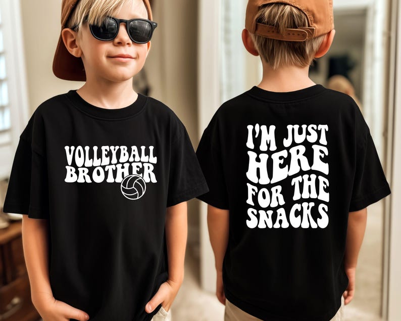 May include: Black t-shirt with white text that reads "Volleyball Brother" and a volleyball graphic on the front. The back of the shirt says "I'm Just Here For The Snacks" in white text. The shirt is worn by a child.