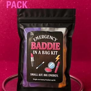 May include: A black pouch labelled "Emergency Baddie in a Bag Kit" with lipstick, a cotton bud, compact, and a lightning bolt graphic. The text "Mini Essentials Pack" is above the pouch. The kit is on a silver tray.