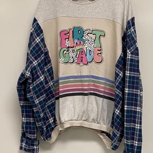 May include: A gray sweatshirt with a plaid shirt sewn onto the sleeves. The sweatshirt has a graphic print that says "FIRST GRADE" in colorful letters with a smiley face and flowers.