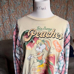 May include: A cream-colored long-sleeved shirt featuring Lola Bunny from Looney Tunes with the word "Peaches" and the phrase "Always Fresh". The sleeves are made from a red, white, and blue plaid pattern.