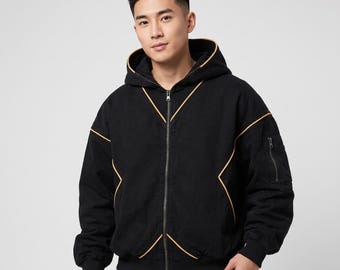 May include: A black zip-up hooded jacket featuring a prominent orange piping detail that forms an X-shape across the front and along the hood. The jacket includes a full-length front zipper, ribbed cuffs, and a small zippered utility pocket on the sleeve.