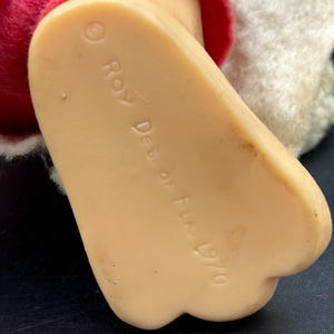 Op de afbeelding: Close-up van een vintage poppenvoet, gemaakt van een lichtbruin materiaal. De voet is licht gebogen met een afgeronde hiel en drie tenen. De tekst "Roy Des. of Fla. 1970" is op de zool gedrukt.