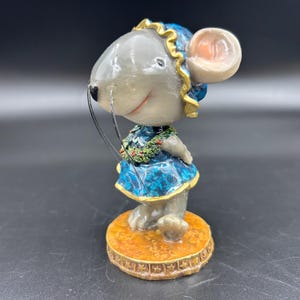 May include: A whimsical figurine of a mouse wearing a blue dress with gold trim and a floral garland. The mouse has a gray body, a pink ear, and long whiskers. It stands on a round, gold-colored base.