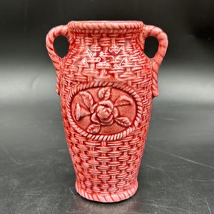 Può includere: Un vaso in ceramica color rosso corallo caratterizzato da un motivo a intreccio di cesto su tutta la superficie. Il vaso presenta due manici laterali e un medaglione ovale centrale con un rilievo di una rosa in fiore con foglie e steli.