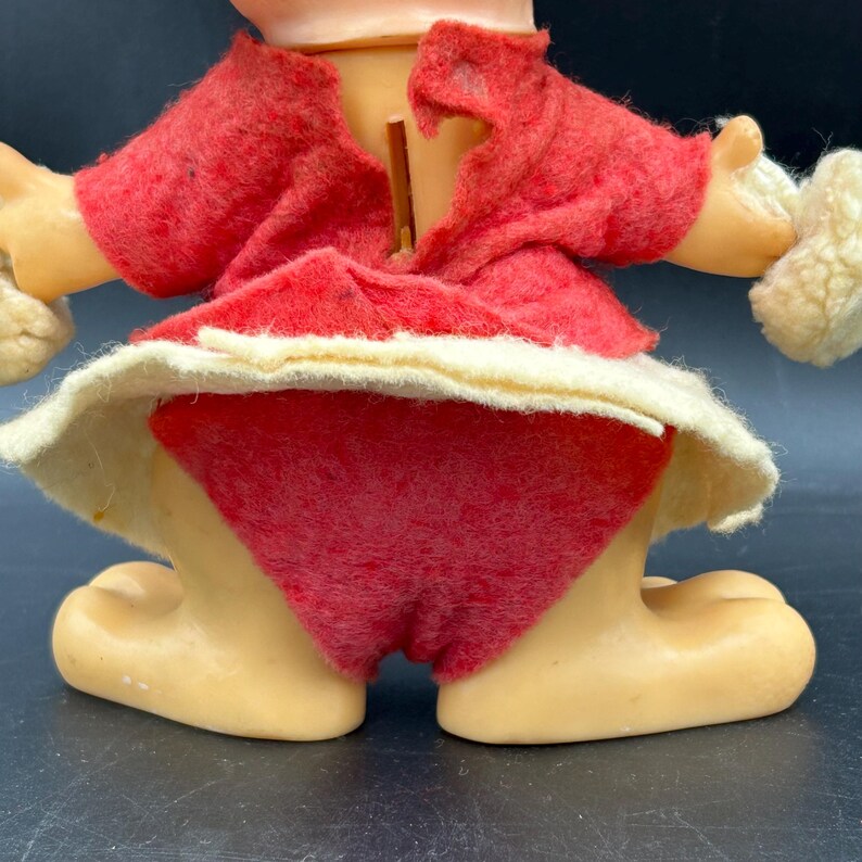 Op de afbeelding: Vintage speelgoedfiguur met een rode vilten outfit en een witte rand. De figuur heeft een lichte huidskleur en houdt twee witte, pluizige objecten vast. Het ontwerp van het speelgoed doet denken aan een klassiek kerstpersonage.