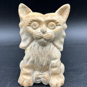 May include: A carved stone cat figurine in shades of tan and beige. The cat is sitting upright with its front paws visible. The cat has detailed facial features, including large eyes and pointed ears. The sculpture has a textured surface.