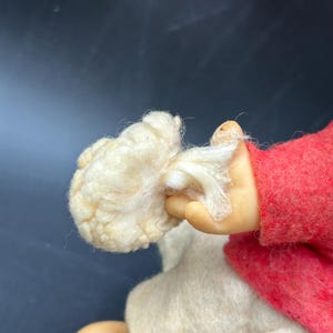 Op de afbeelding: Een close-up van een pop met een cr&egrave;mekleurige wollige baard en een rode vilten jas. De hand van de pop, met een lichte huidskleur, houdt de baard vast. Het lichaam van de pop is gemaakt van een zacht, wit materiaal.