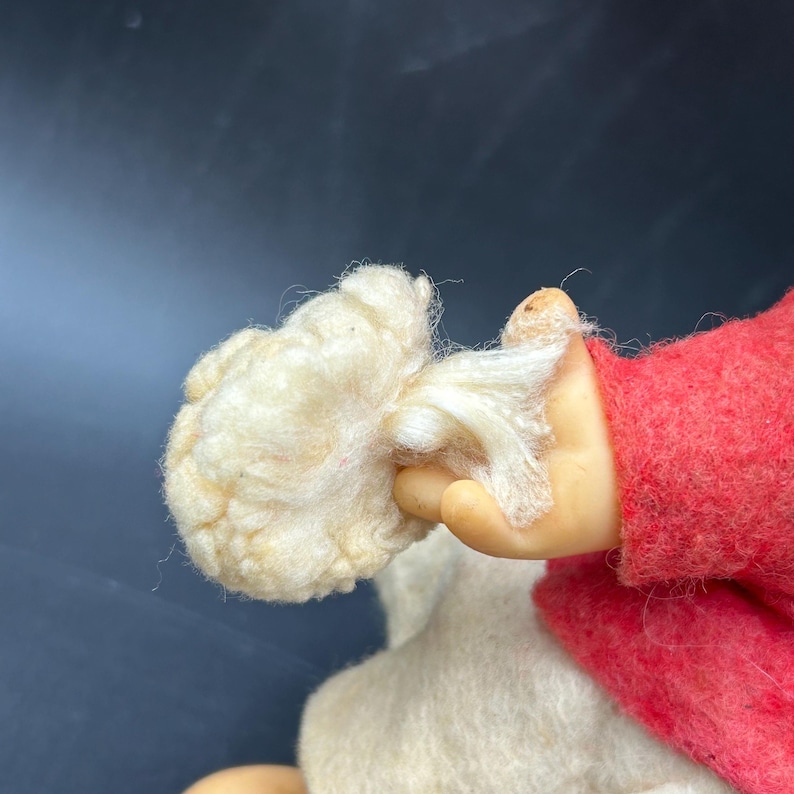 Op de afbeelding: Een close-up van een pop met een cr&egrave;mekleurige wollige baard en een rode vilten jas. De hand van de pop, met een lichte huidskleur, houdt de baard vast. Het lichaam van de pop is gemaakt van een zacht, wit materiaal.