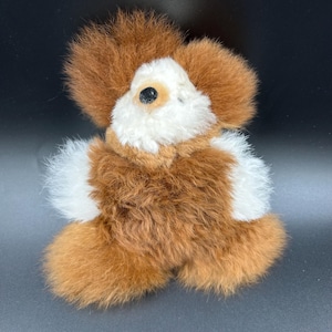 May include: A plush dog toy with brown and white fur. The toy has a fluffy appearance with a brown head and ears, white face and chest, and brown body. The dog has a black eye and is sitting upright.