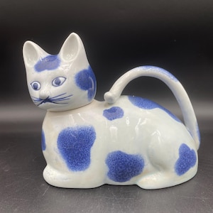 May include: A white ceramic teapot shaped like a cat with blue and white spots. The teapot has a lid and a handle that is shaped like a cat's tail.