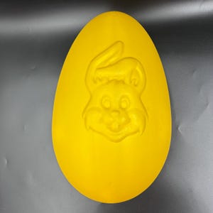 May include: A bright yellow, egg-shaped decorative item with a carved bunny face. The bunny has large eyes, a smiling mouth, and long ears. The item is likely used for Easter decorations.