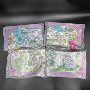 May include: Four rectangular plastic-wrapped maps of a theme park, each featuring colorful illustrations of park attractions, pathways, and green spaces. The maps include labels such as Frontierland, Fantasyland, and Tomorrowland, with a small toy figure.