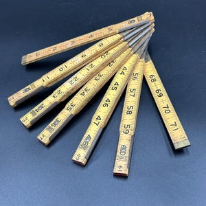 May include: A vintage wooden folding ruler, featuring a yellowed finish with black measurement markings in inches. The ruler is partially open, displaying numbers up to 71 inches. The metal joints show signs of age, adding to its antique charm.