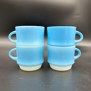 May include: Four vintage light blue stackable mugs with white bases and curved handles. The mugs are arranged in two stacks of two, showcasing their unique design and color contrast. The mugs are made of glass.