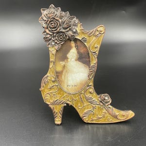 May include: A decorative picture frame shaped like a high-heeled boot. The frame is gold with brown accents and features a vintage portrait. The frame is 2.5" x 3.5" and adorned with roses and rhinestones.