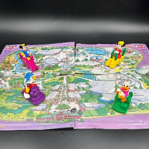 May include: A board game featuring a colourful map of a theme park with four plastic character figurines. The figures include Mickey Mouse on a pink slide, Minnie Mouse on a yellow base, Donald Duck on a green base, and Goofy on a purple base.