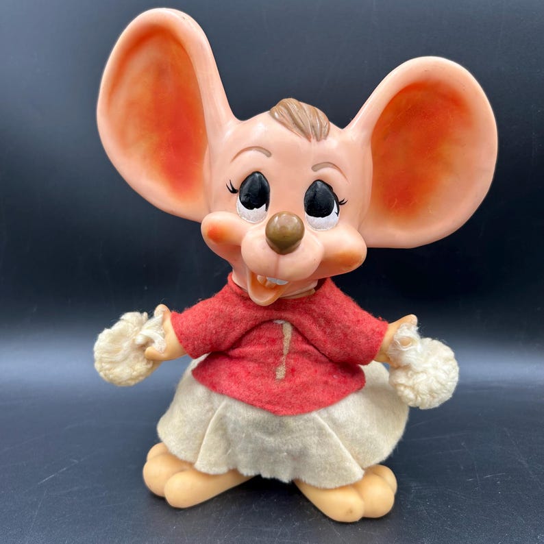 Op de afbeelding: Vintage beeldje van een muisfiguur met grote oren, gekleed in een rode top en een cr&egrave;mekleurige rok. De muis heeft een lachende uitdrukking, zwarte ogen en beige poten. Een verzamelobject.