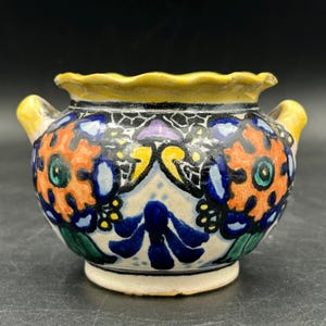 May include: A small, hand-painted ceramic vase with a yellow rim and handles. The vase features a colorful floral design in orange, blue, green, and purple against a cream background. The vase has a scalloped rim and is likely a decorative item.
