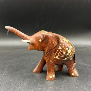 May include: A carved wooden elephant figurine with a raised trunk and ivory-colored tusks. The elephant is brown with decorative white inlay on its back and feet. The figurine is approximately 5 inches long.