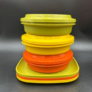 Peut inclure: Une pile de quatre récipients alimentaires ronds en plastique avec couvercles. Le récipient supérieur est vert olive, le deuxième est jaune, le troisième est orange et la base est une assiette carrée vert olive. Chaque récipient est lisse.