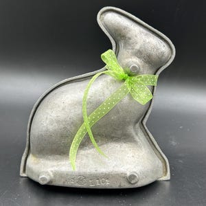 May include: A vintage metal bunny-shaped mold, perfect for baking. The mold is a dull silver color and features a green ribbon tied around the neck. The mold has a detailed bunny shape with a textured surface and is ideal for Easter baking.