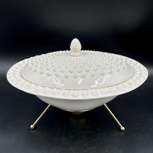 May include: A white ceramic serving bowl with a matching lid, both featuring a raised hobnail texture. The bowl is supported by three thin, gold-toned metal legs that extend outward from the base, providing a raised stance for the dish.