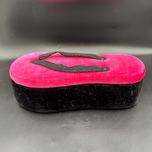 May include: A rectangular jewelry box with a black base and a vibrant pink top. The top is adorned with a black handle. The box is made of a soft, velvety material, creating a luxurious appearance. The design is simple yet elegant.