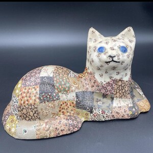 May include: A white ceramic cat figurine with blue eyes. The cat is lying down and is covered in a patchwork of colorful fabric.