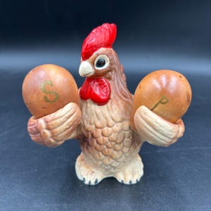 May include: A ceramic rooster-shaped salt and pepper shaker set. The rooster has a red comb and wattle, brown feathers, and holds two brown egg-shaped shakers, one marked with an "S" and the other with a "P".