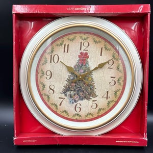 May include: A 15.75-inch round wall clock with a white and gold frame. The clock face features a beige crackle finish, a central Christmas tree illustration with a red bow, and black numerals. The text 15.75" caroling clock is printed on the red box.