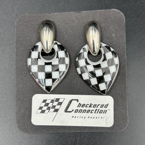 May include: A pair of vintage earrings with a silver-tone top and a teardrop-shaped checkered design in black and white. The earrings are displayed on a black background with a label that reads "Checkered Connection Racing Apparel."