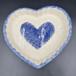 May include: Heart-shaped ceramic serving dish with a cream-colored interior and a blue speckled heart design in the center. The rim and the heart's outline are also speckled blue. A charming piece for serving or display.