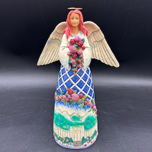 May include: A decorative angel figurine with pink hair, holding a bouquet of pink roses. The angel wears a white top and a blue dress with a diamond pattern. The base features a green landscape and a white picket fence.