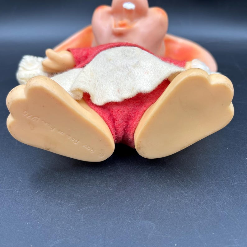 Op de afbeelding: Een vintage speelgoedfiguur van een stripfiguur, met een roze gezicht en oren, een rode outfit en cr&egrave;mekleurige afwerking. De grote, beige voeten van de figuur zijn zichtbaar, met de tekst "Roy Des. Inc. 1970".