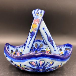 May include: A small, decorative ceramic basket with a handle. The basket is primarily white with blue accents and floral designs. The handle is arched and decorated with colorful flowers and leaves. The basket has a pierced design around the rim.