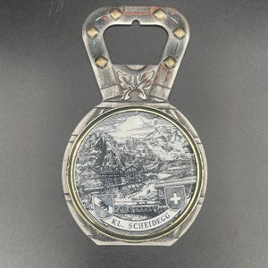 May include: Vintage metal bottle opener with a decorative design. The opener features a detailed black and white illustration of a mountain landscape with buildings, a gold border, and the text "KL. SCHEIDEGG".