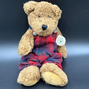 May include: A plush teddy bear with brown fur, wearing red and blue plaid overalls. The bear has a black nose and small blue eyes. A tag that says "Boyds Bears Bearwear" is attached to the bear.