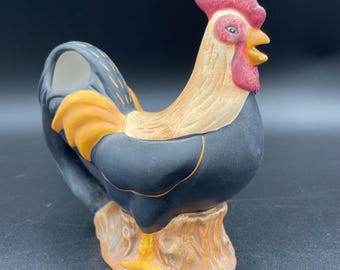 May include: A black, yellow, and red rooster creamer with a brown base. The rooster is standing on a tree stump.